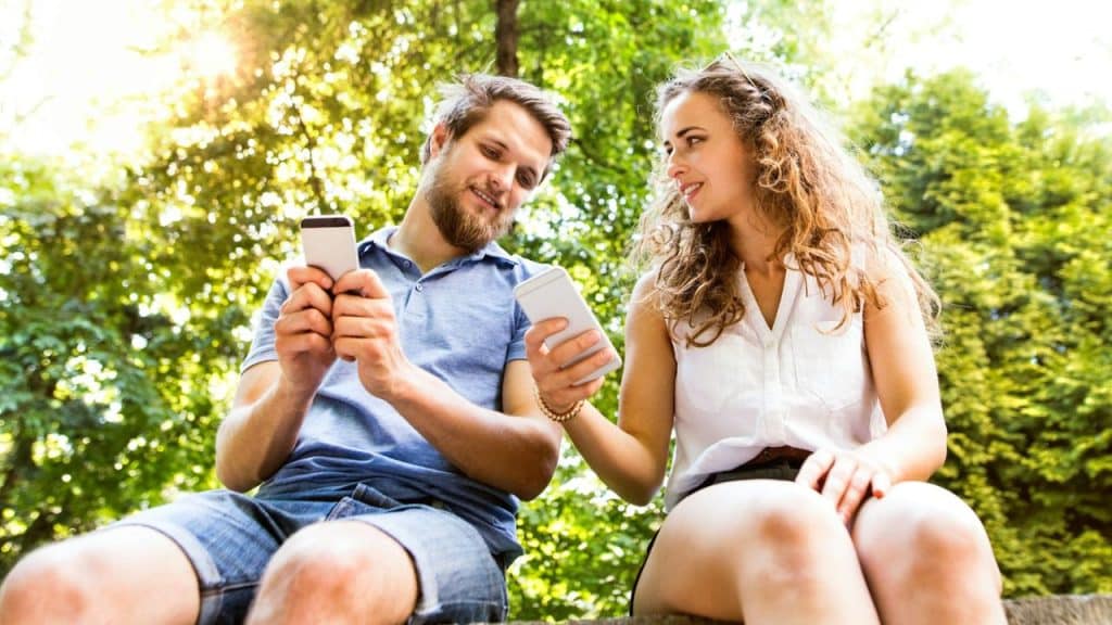 A man and woman sit outside under trees while looking at their mobile phones.