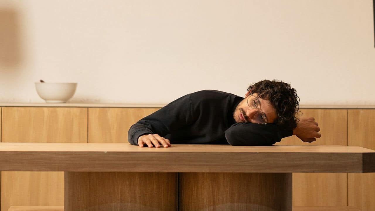 A man wearing glasses rests his head on his arm atop a wooden table.