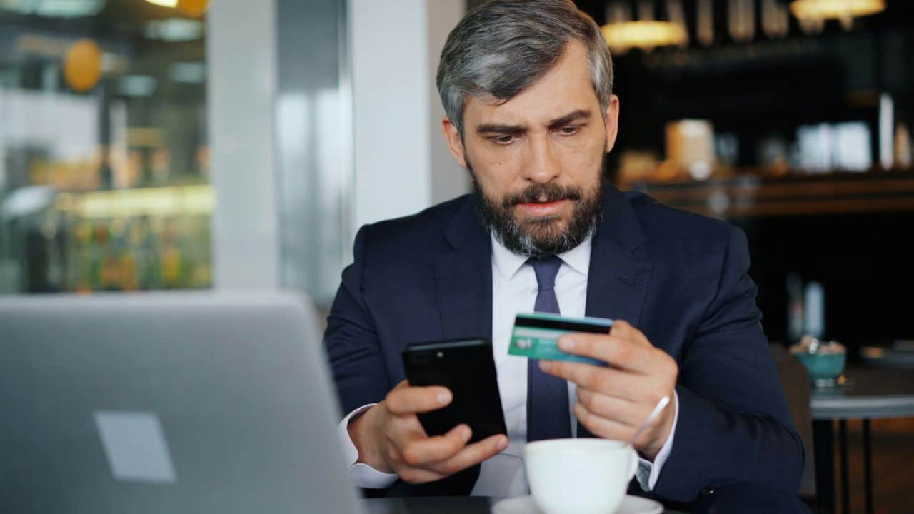 A man in a suit holds a credit card and a smartphone while sitting at a table.