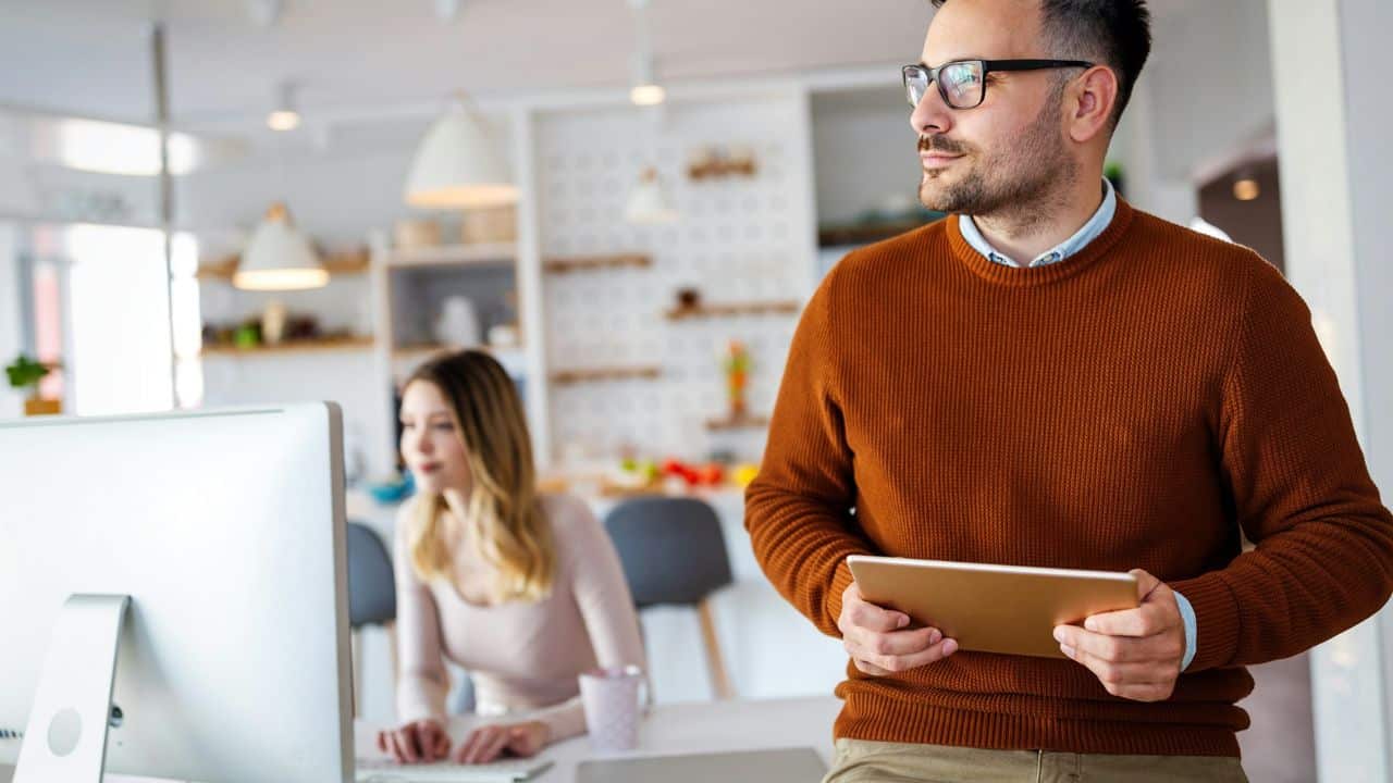 A man stands holding a tablet while a woman works at a computer behind him.