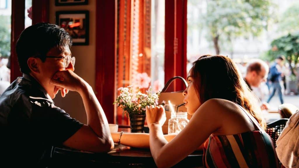 A man and woman sit at a small table inside a cafe with bright sunlight.