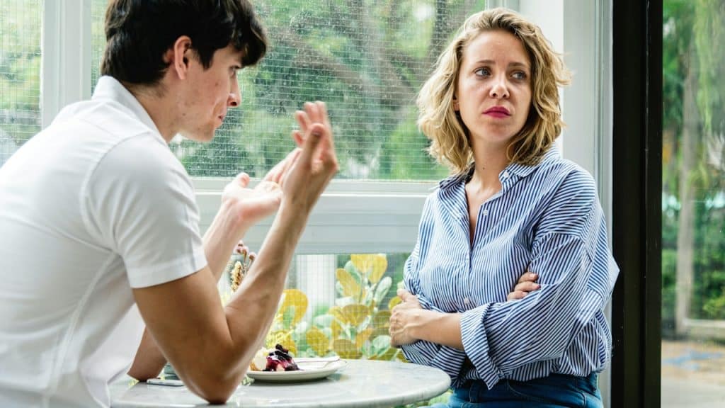 A man trying to explain something to a woman who looks away while folding her arms across her chest as they sit at a table.
