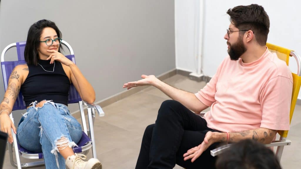A bearded man talking to a woman wearing glasses and gesturing towards her while sitting on folding chairs.