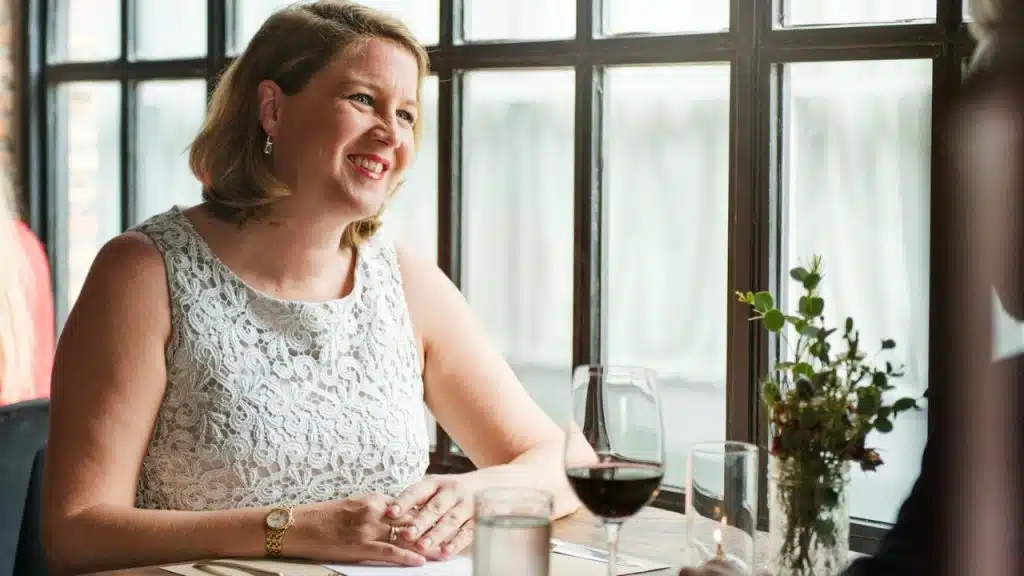 A woman in a white lace dress smiles while sitting at a table with wine.