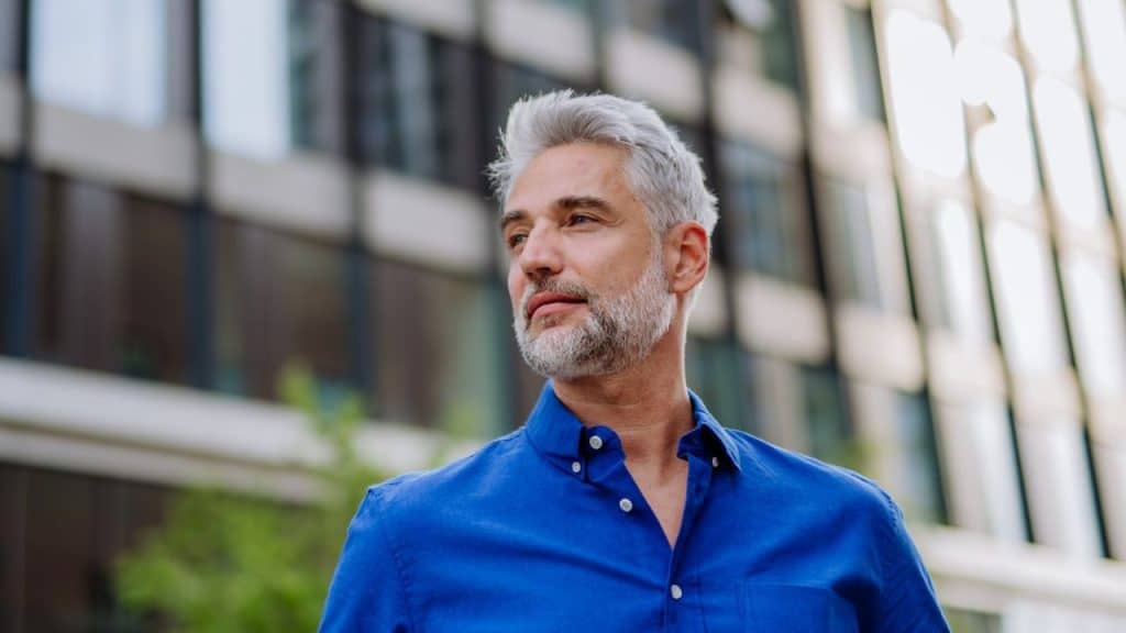 A gray-haired man in a blue shirt looks off to the side against a blurred building.