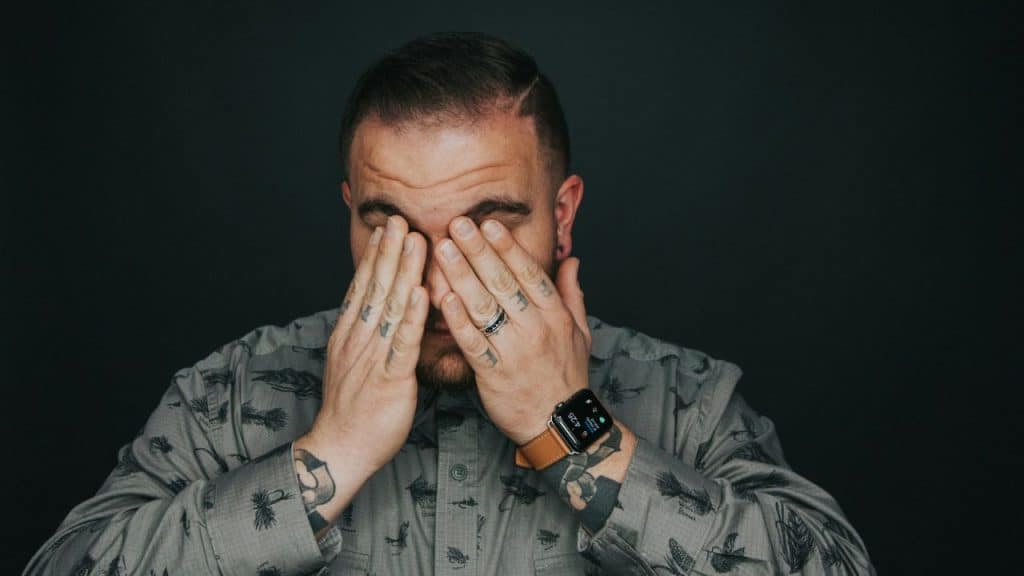 A man with tattoos on his hands and arms covers his eyes with his fingers while looking down.