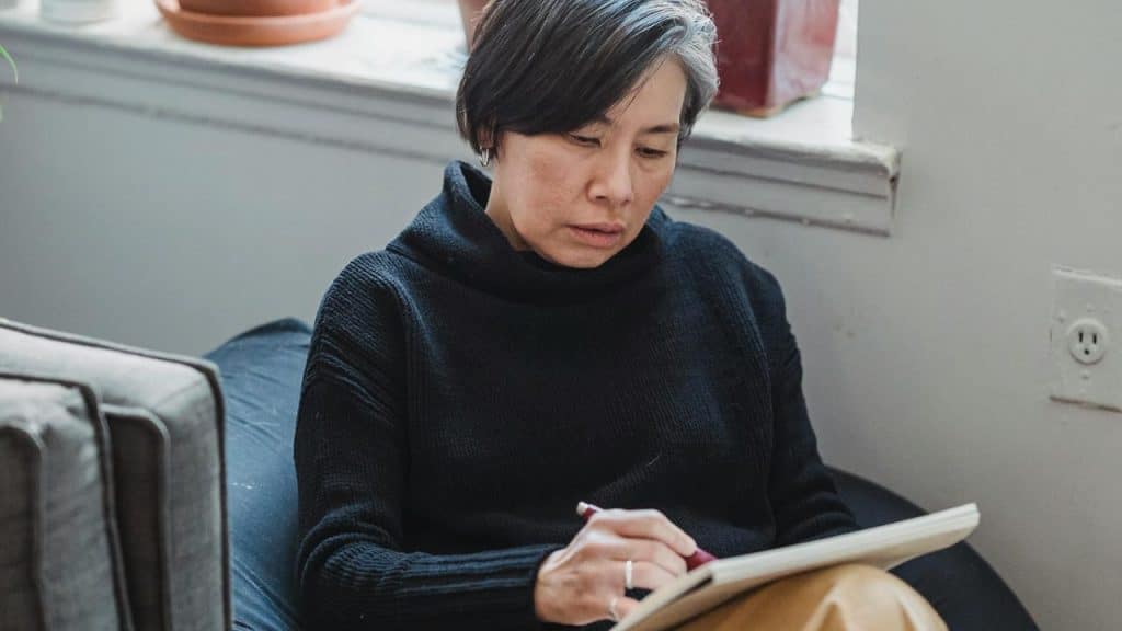 A woman sitting on a couch, writing in a notebook near a window.