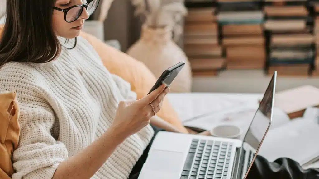 A woman wearing glasses looks at her phone while sitting with a laptop.