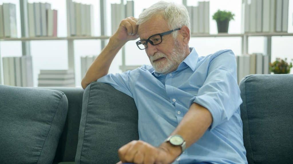An older man with grey hair and glasses sits on a sofa looking downward pensively.