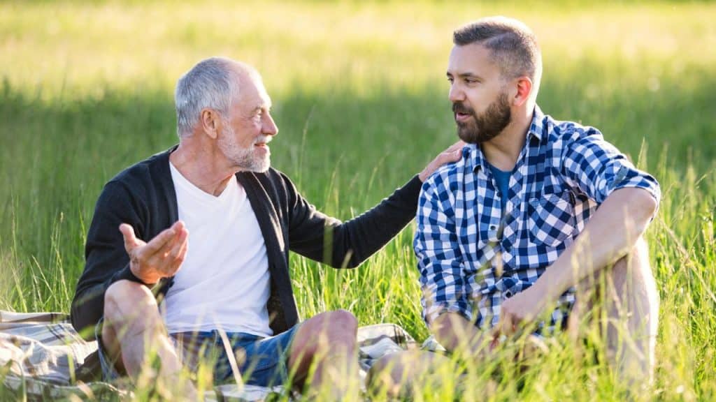 An older man and a younger man sit on a blanket in a grassy field talking.