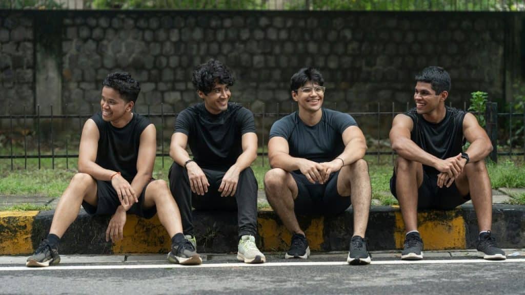 Four men in athletic wear sit together on a curb, smiling and talking.