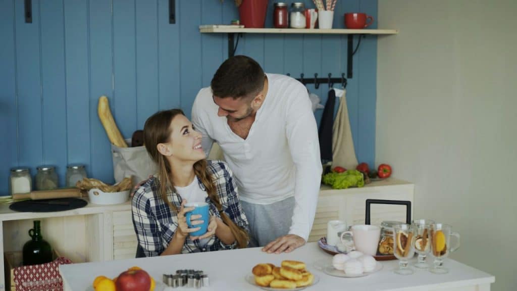 A man in a white shirt and a woman in a plaid shirt smile together in a kitchen.