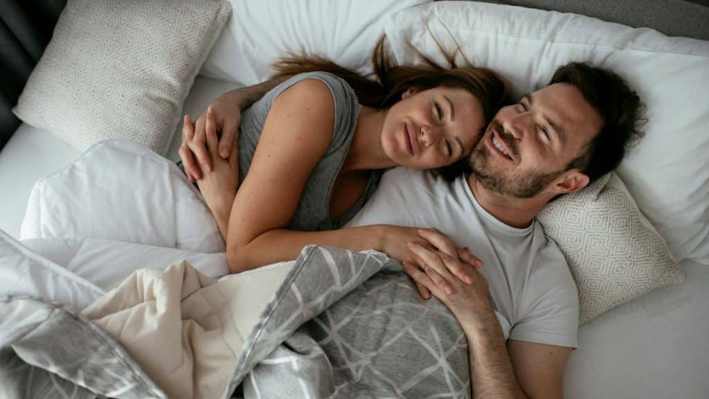 A man and woman lie in bed, smiling and holding hands under a grey patterned blanket.