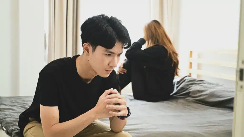 A man sits on a bed, looking down, while a woman sits turned away in the background.