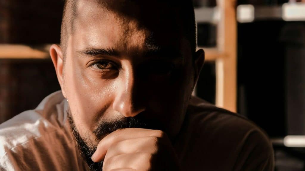 A close-up of a man with a beard looking directly at the camera with a hand on his chin.