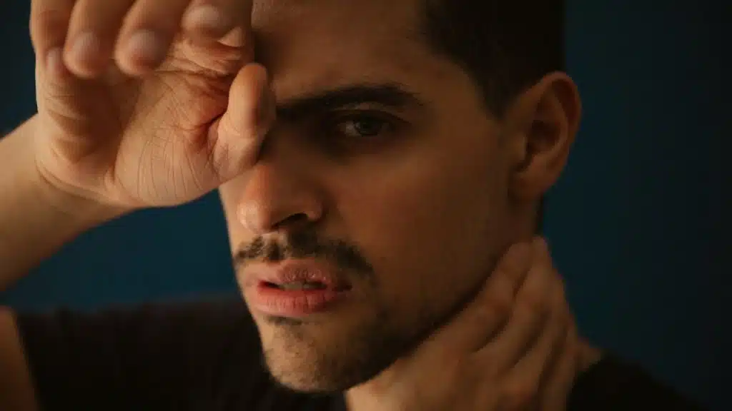 A man with a mustache covers one eye with his hand while looking forward.