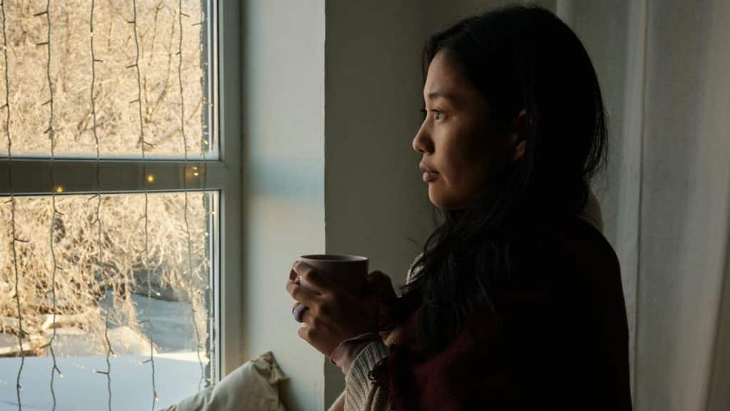 A woman holds a mug while looking out a window decorated with hanging string lights.
