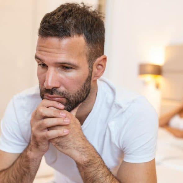A man sits on a bed looking thoughtful while a woman sits blurred in the background.