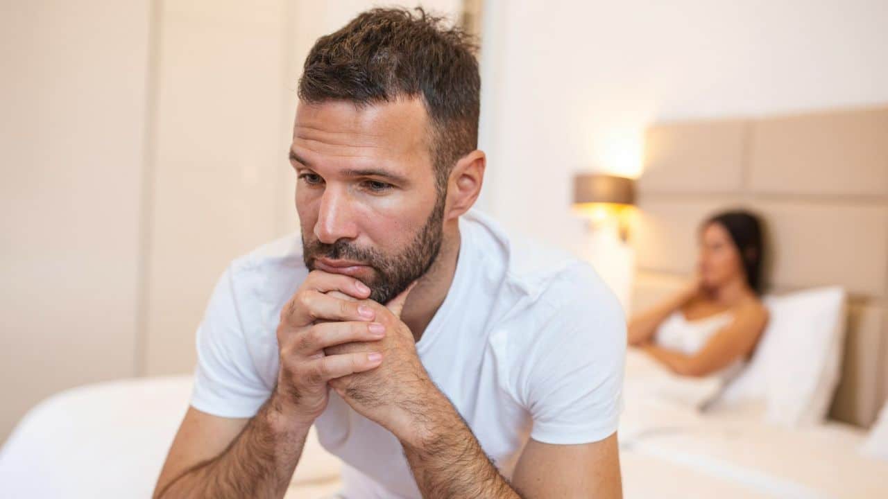 A man sits on a bed looking thoughtful while a woman sits blurred in the background.
