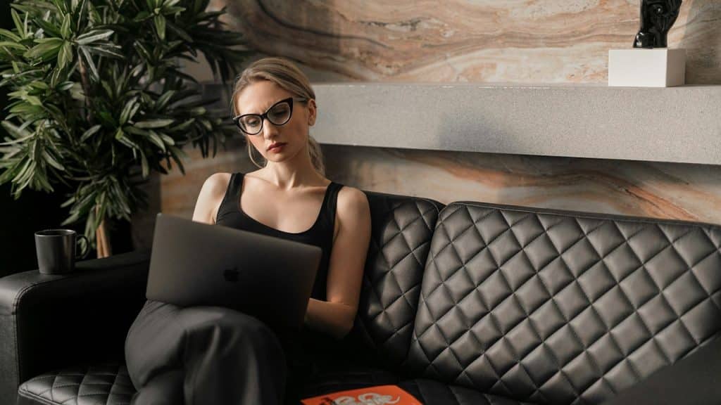 A woman wearing glasses working on her laptop while sitting on a couch.