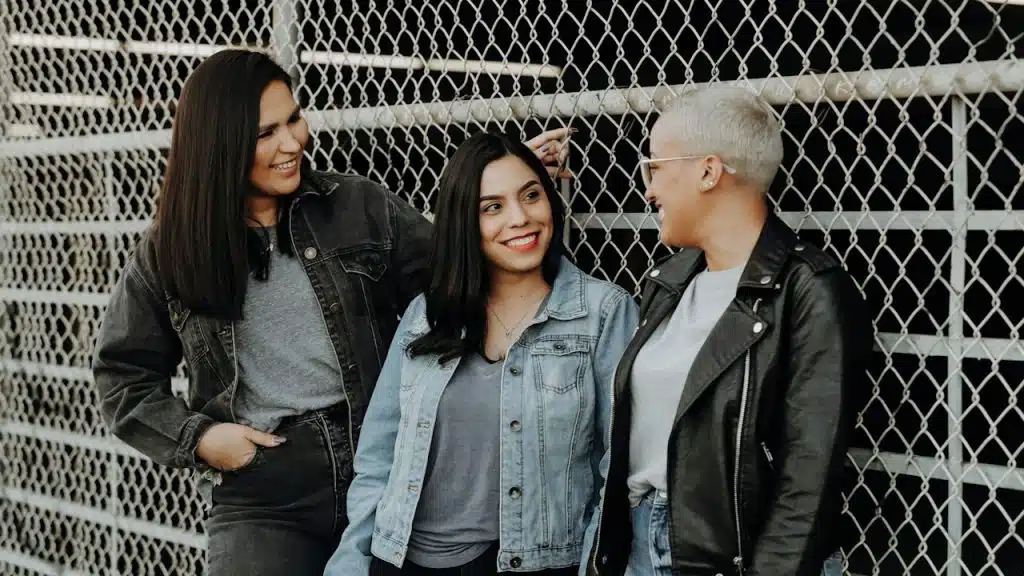 A trio of friends talking and smiling while leaning against a fence.