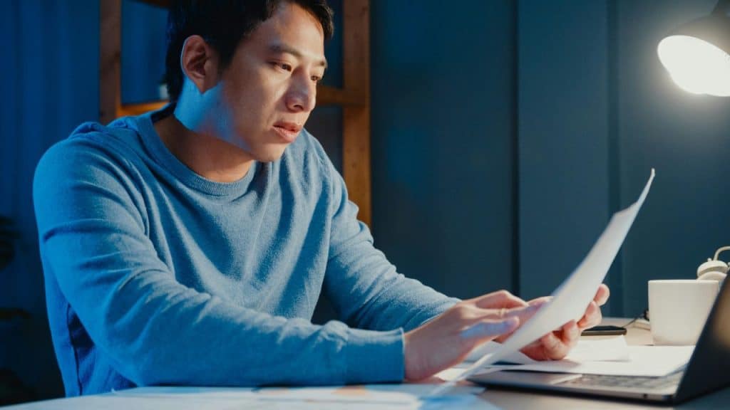 A man in a blue sweater reviews paperwork at a desk under a lamp.