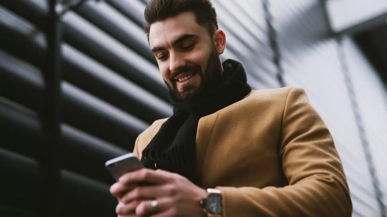 A man wearing a brown coat smiling while texting outdoors.