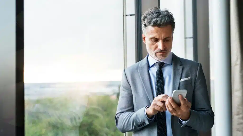 A man in a grey suit looks down while using his smartphone near a window.