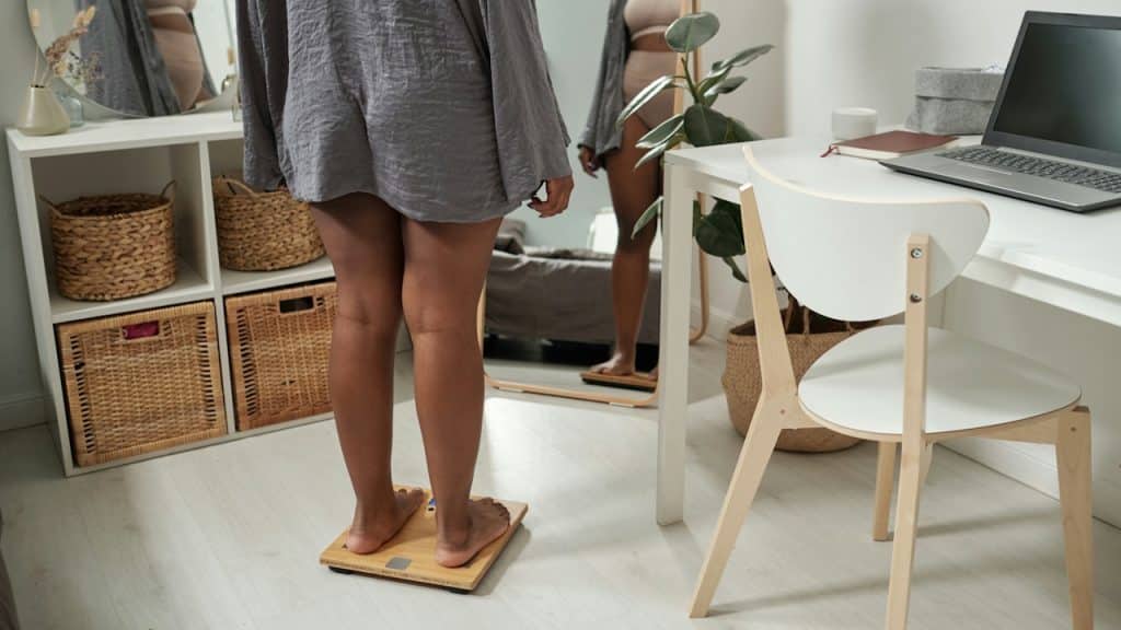 A woman stands on a wooden digital scale in a bright room, looking at her reflection in a full-length mirror.