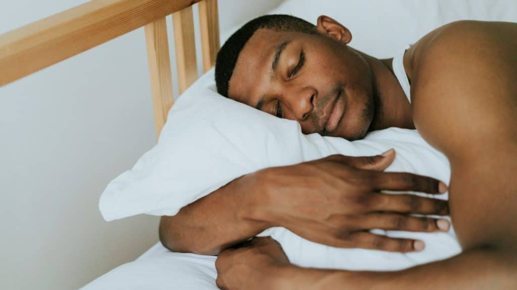 A man with his eyes closed rests his head on a white pillow while hugging it.
