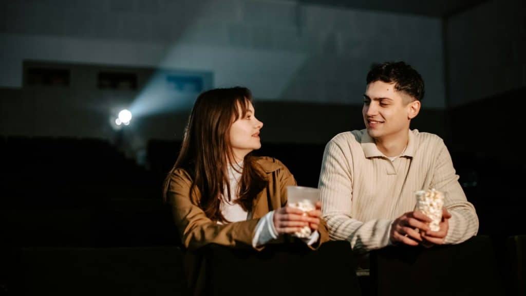 A man and woman sit in a theater holding popcorn and looking at each other.