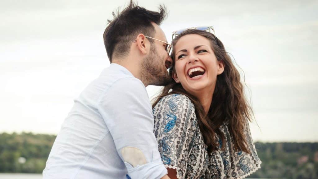 A man whispers to a woman who is laughing heartily outdoors.