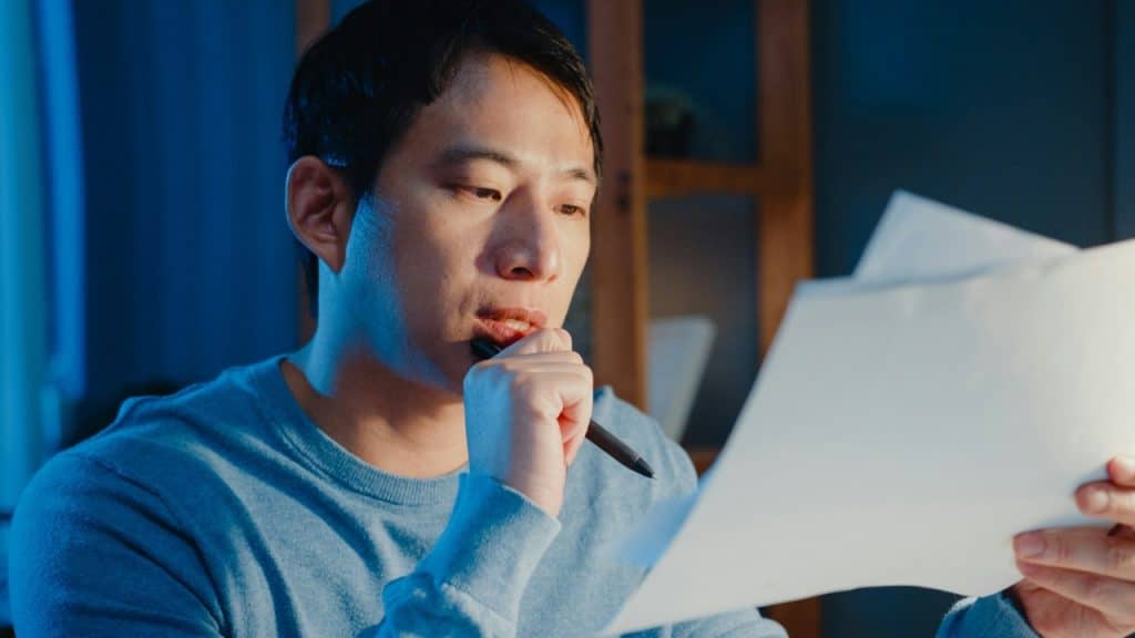 A man in a blue sweater holds papers and rests a pen against his chin.