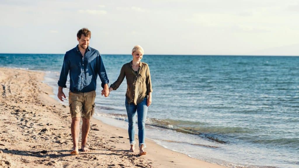 A man and woman hold hands while walking along a sandy beach near the ocean.