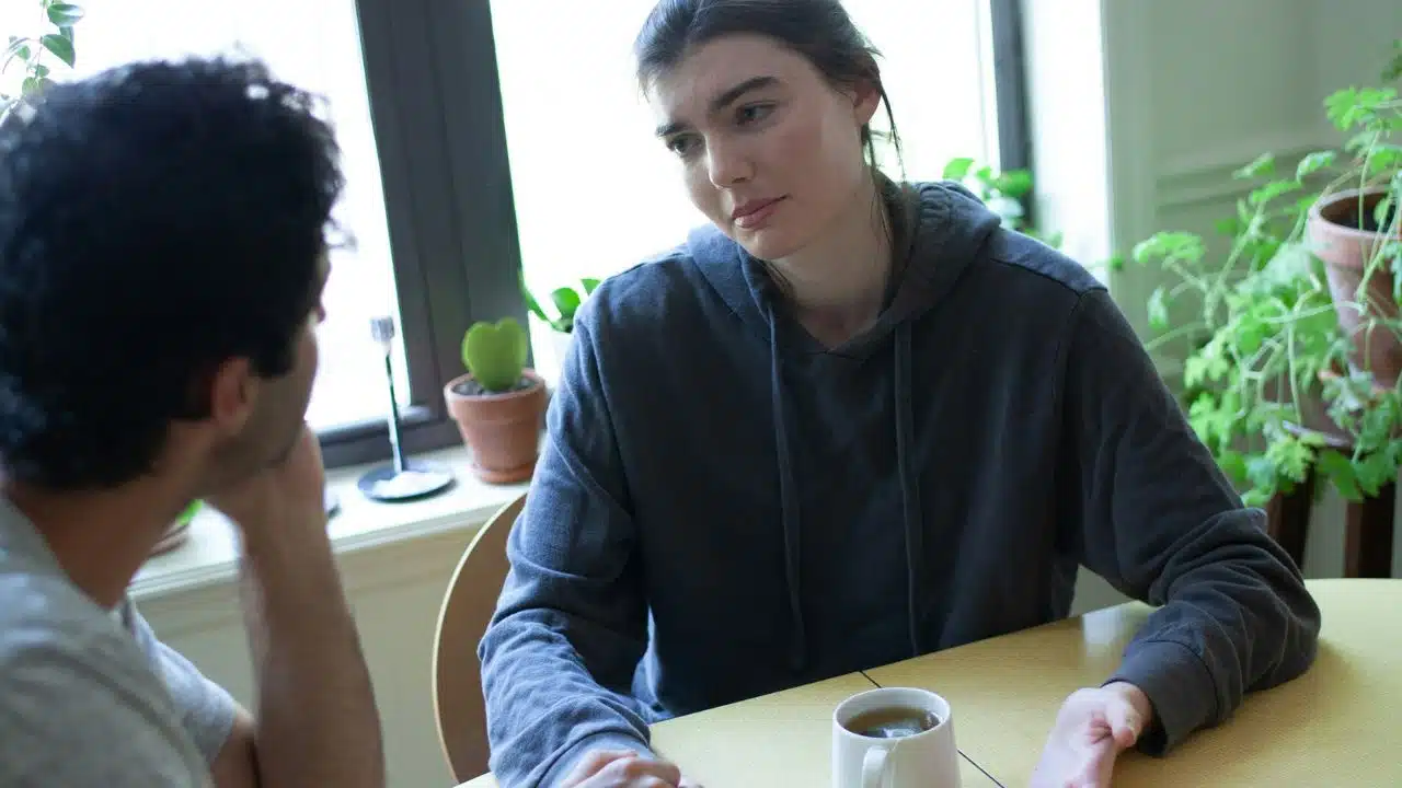 A woman in a grey hoodie sits at a table listening to a man speaking.