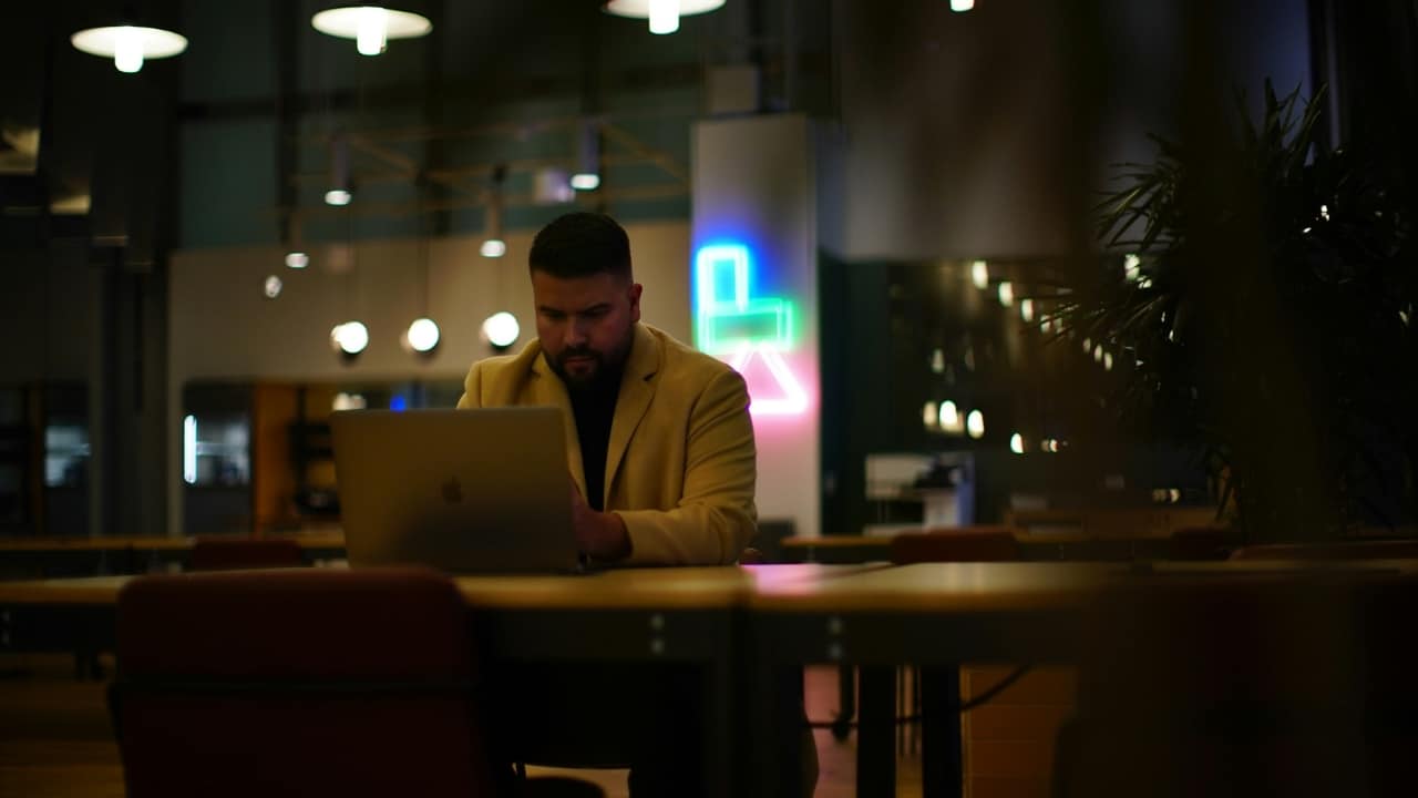 A man in a tan blazer works on a laptop in a dimly lit office space.