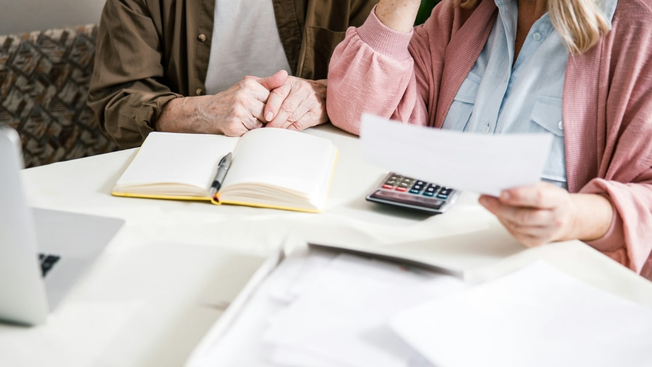 Two people sit at a table with an open notebook, a calculator, and various papers.