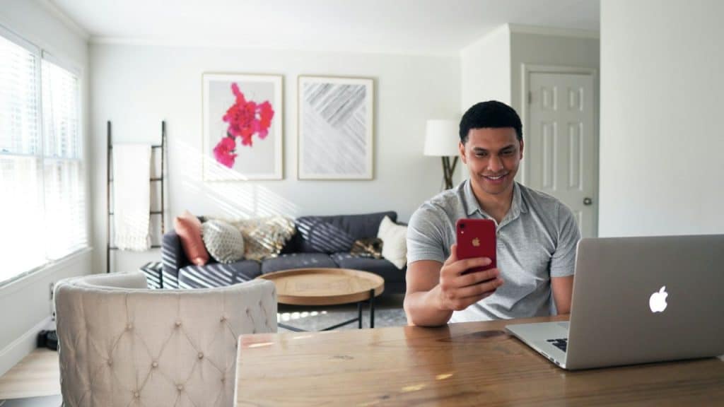 A man sits at a wooden table smiling while looking at a red smartphone and laptop.