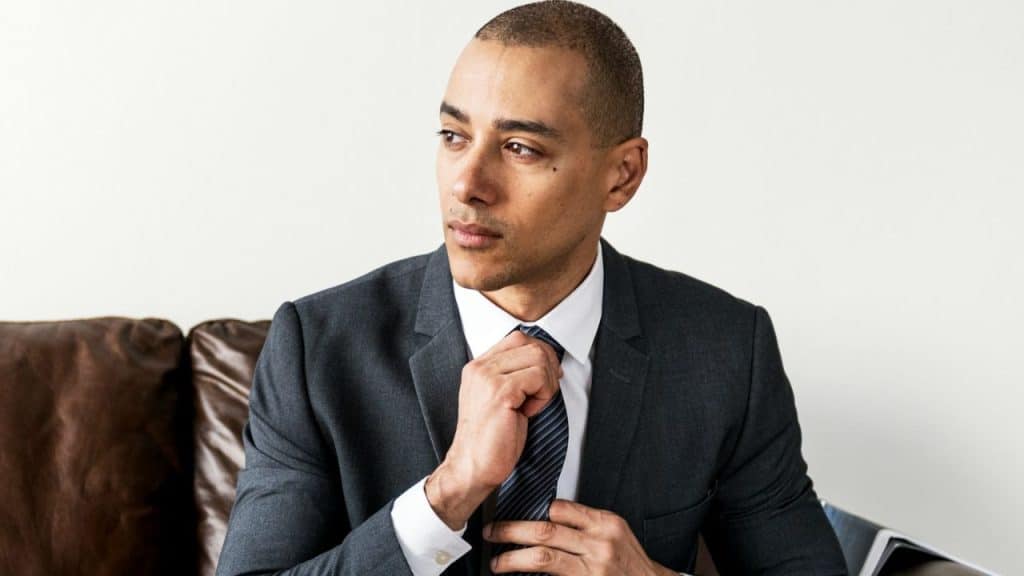 A man in a dark suit and white shirt adjusts his tie while looking off-camera.