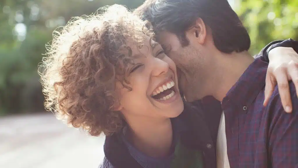 A man whispering into a laughing woman’s ear outdoors.