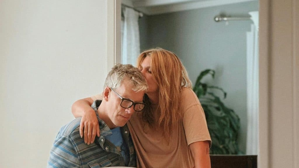 A woman hugging and kissing a man while he writes at a table indoors.