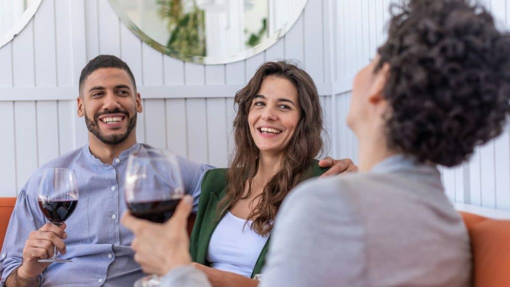 A smiling man and woman sit with wine glasses while looking at a third person.