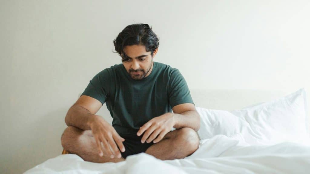 A man sits cross-legged on a white bed looking down with a somber expression.