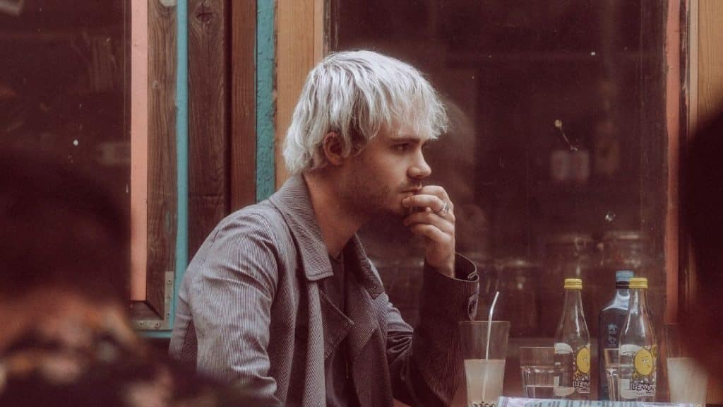 A man with light-colored hair sits at a table outdoors and rests his chin on his hand.