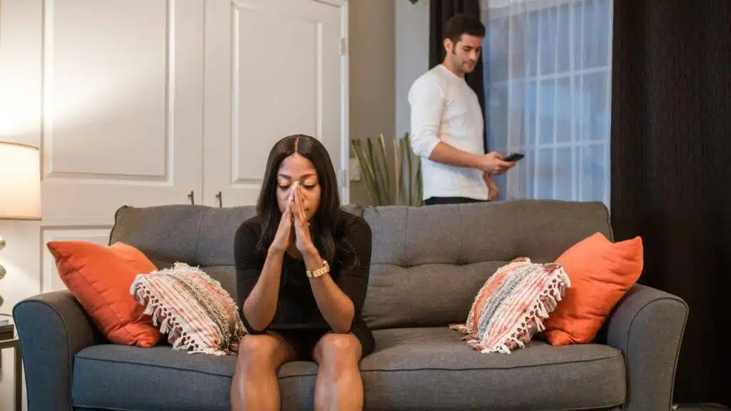 A woman sits with her head in her hands while a man stands in the background.