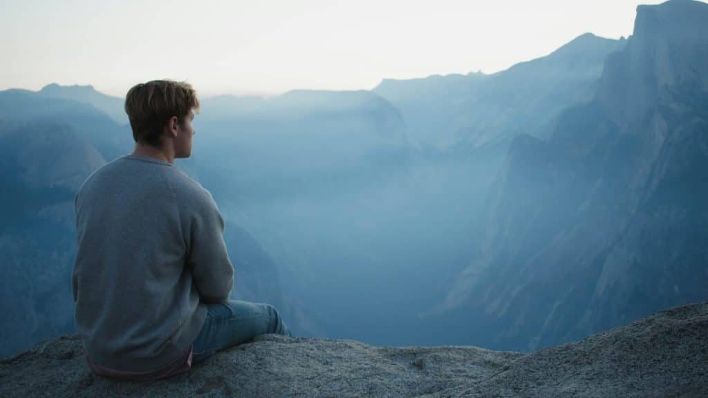 A man sits on a rocky cliff edge looking out over a misty mountain valley.