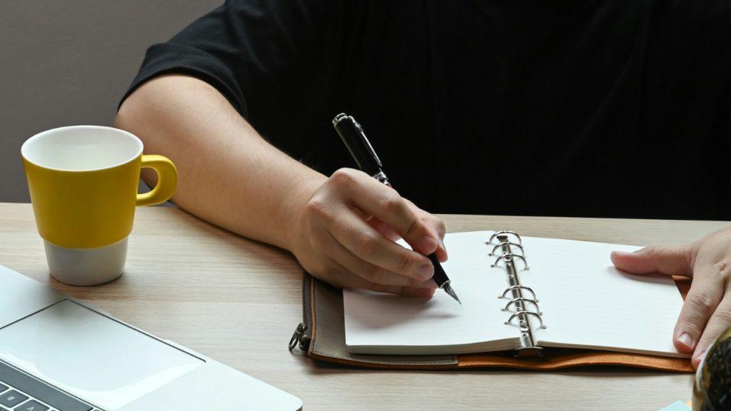 A person's hand writes with a fountain pen in a ringed notebook on a desk.