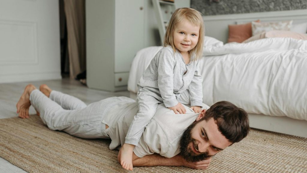 A toddler sitting on a father’s back as he lies on the floor.