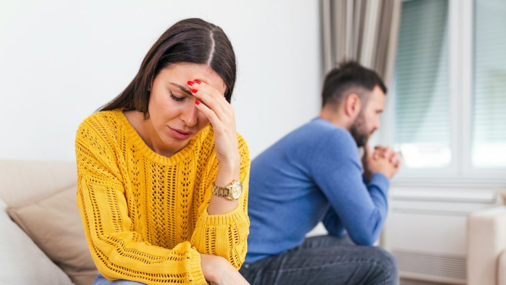 A woman in a yellow sweater holds her forehead while a man sits in the background.