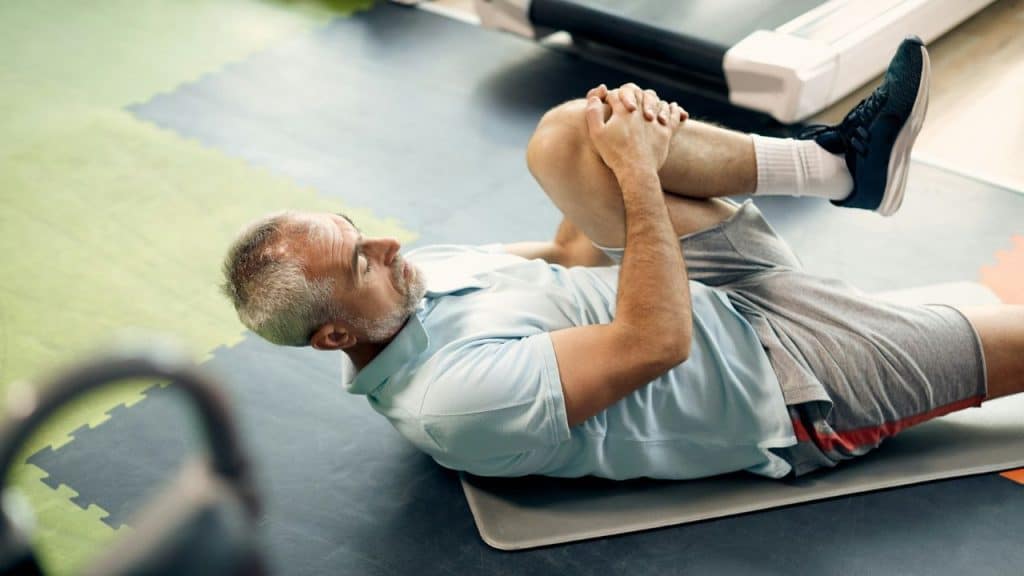 An older man lying on a gym mat pulls one knee toward his chest to stretch.